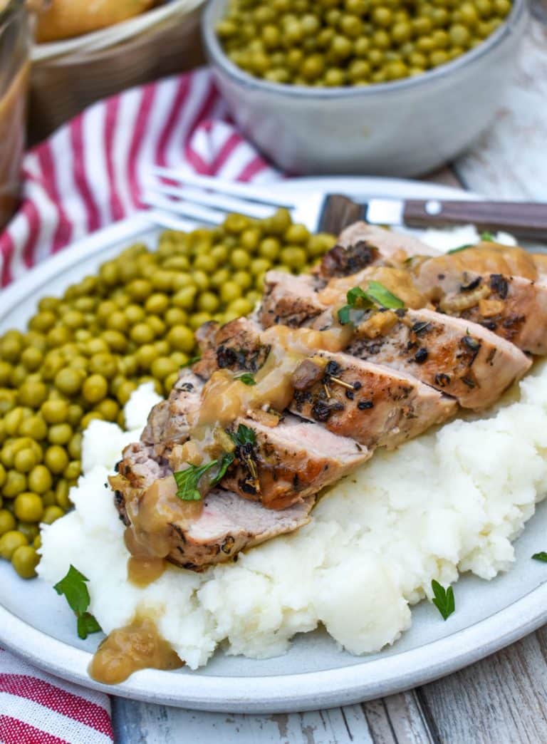 slices of pork tenderloin with apple cider gravy over mashed potatoes next to baby peas on a white plate