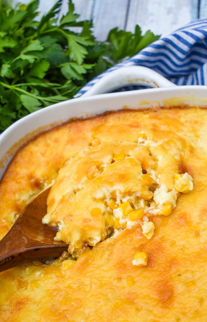 a wooden serving spoon digging into baked corn pudding in a white casserole dish