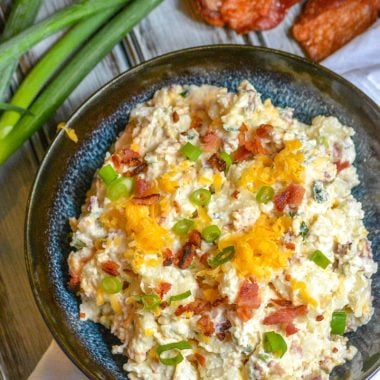 loaded baked potato salad in a blue bowl next to green onions and crispy strips of cooked bacon