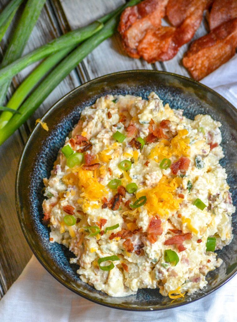 Loaded Baked Potato Salad in a blue ceramic bowl with green onions and crispy bacon in the background