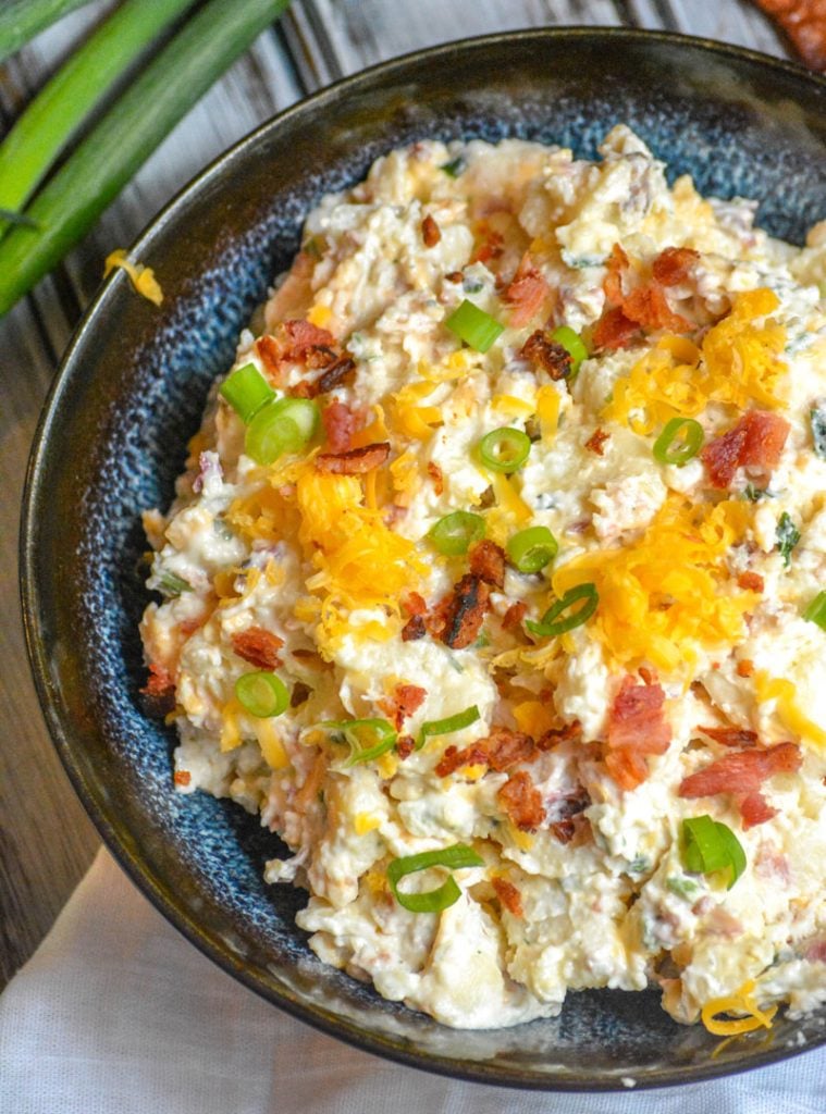 Loaded Baked Potato Salad in a blue bowl, topped with shredded cheese, bacon crumbles, and sliced green onions