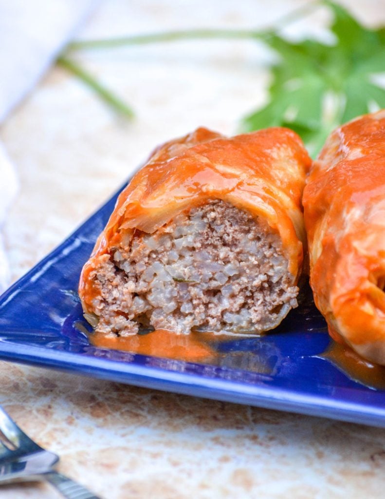 saucy Halupki Stuffed Cabbage Rolls serve on a blue square plate with a sprig of parsley and a silver serving spatula in the background