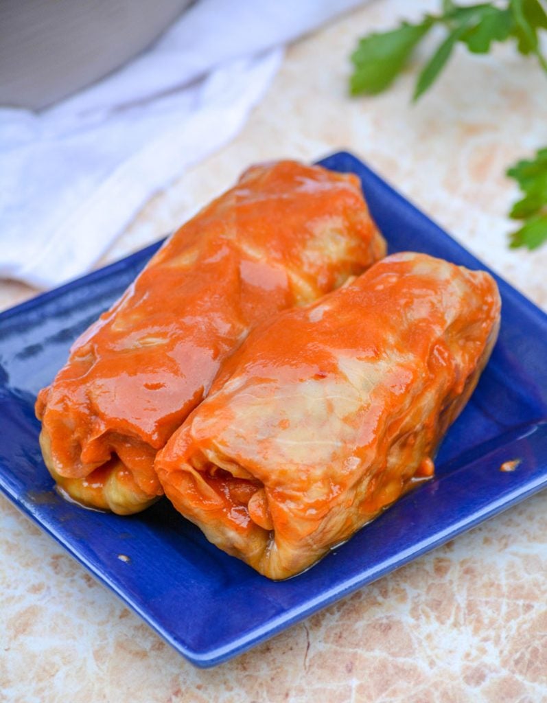 saucy Halupki Stuffed Cabbage Rolls serve on a blue square plate with a sprig of parsley and a silver serving spatula in the background