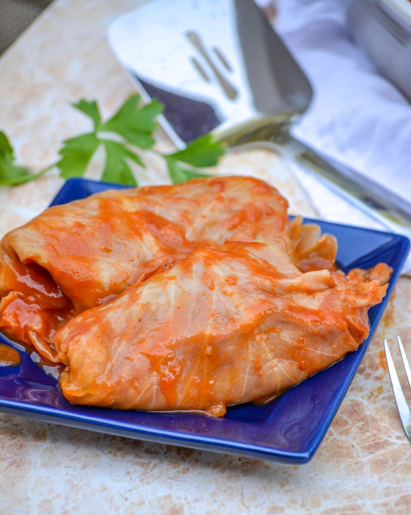 saucy Halupki Stuffed Cabbage Rolls serve on a blue square plate with a sprig of parsley and a silver serving spatula in the background