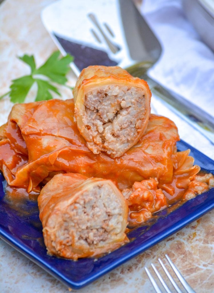 saucy Halupki Stuffed Cabbage Rolls serve on a blue square plate with a sprig of parsley and a silver serving spatula in the background