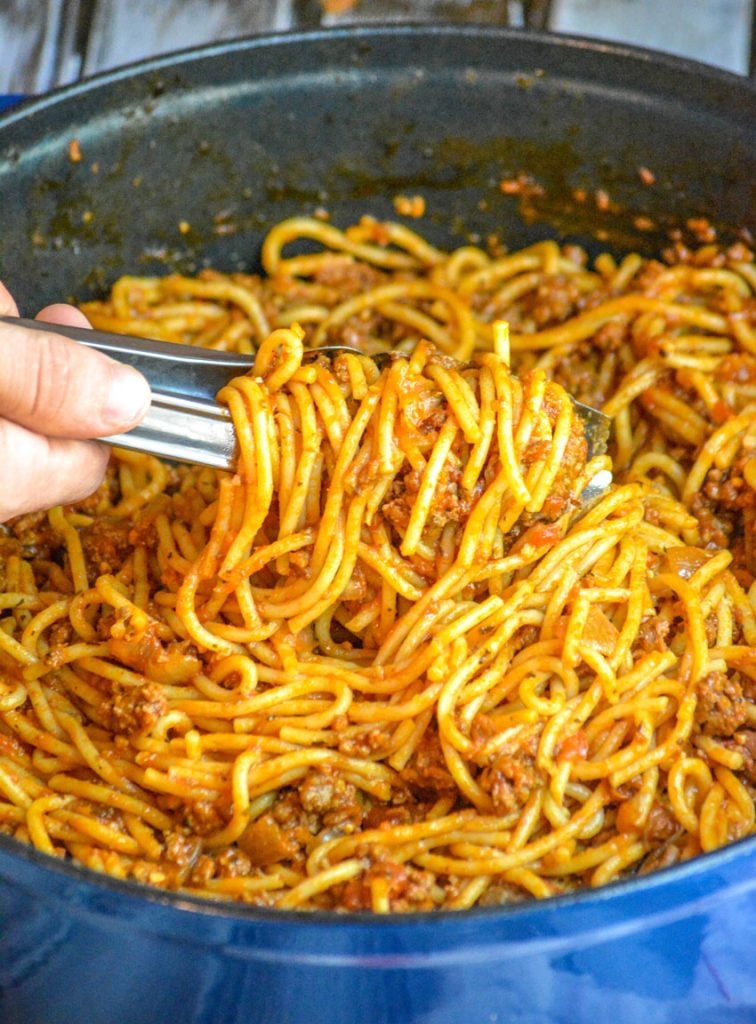 a hand holding tongs scooping up One Pot Spaghetti from an enameled cast iron pot