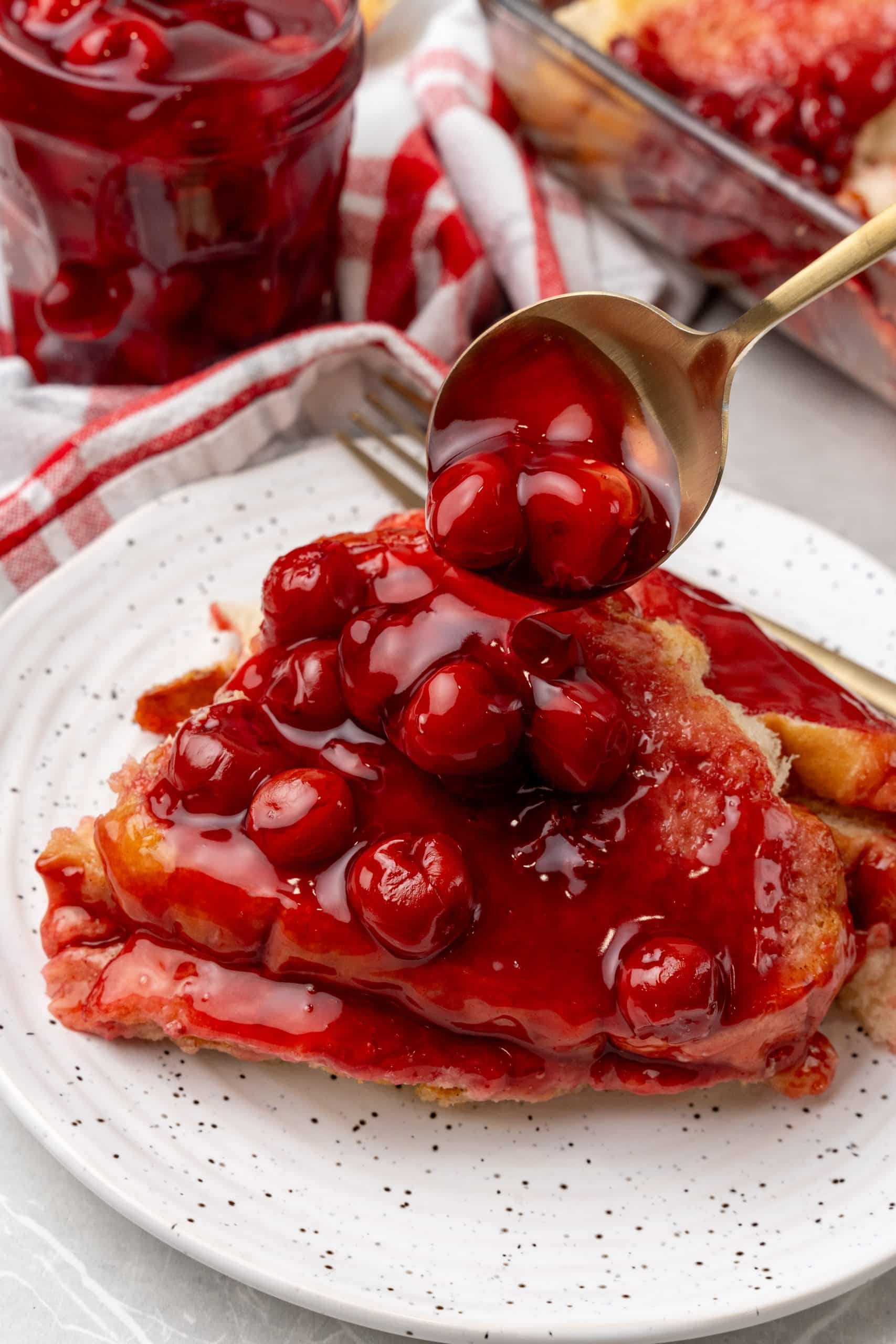 a gold spoon spooning cherry pie filling over a slice of a cherry french toast casserole