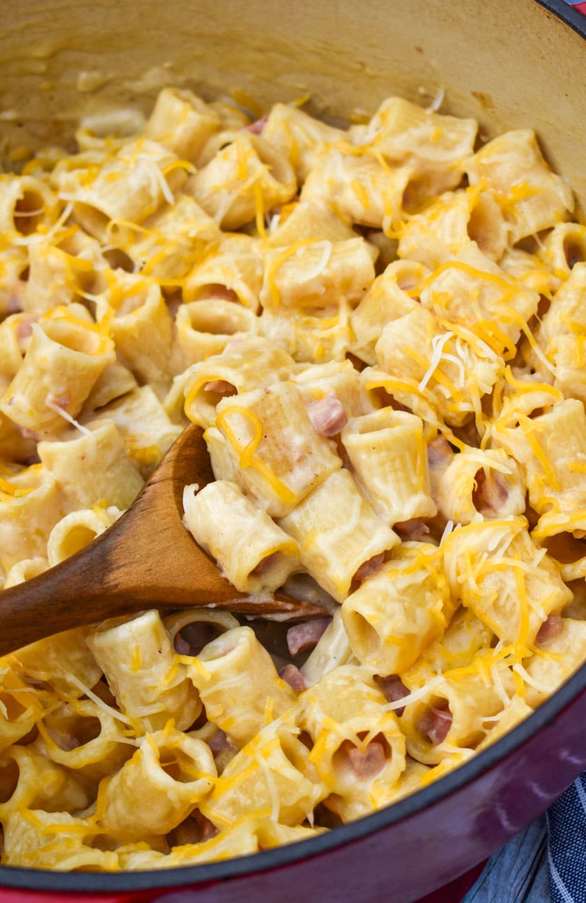 a wooden spoon scooping cheesy riggis pasta with ham out of a large pot