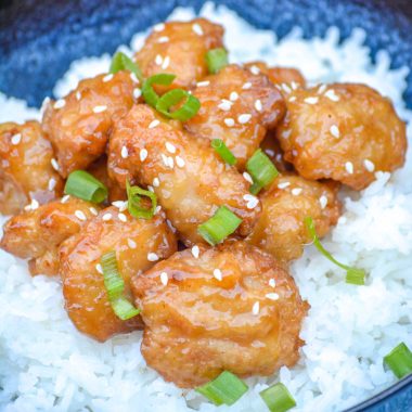 ORANGE CHICKEN TOPPPED WITH SESAME SEEDS AND SLICED GREEN ONIONS OVER COOKED WHITE RICE IN A BLUE BOWL