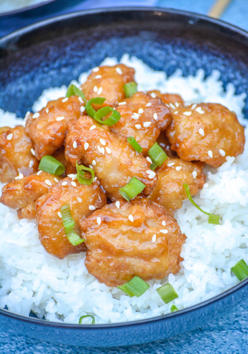 ORANGE CHICKEN TOPPPED WITH SESAME SEEDS AND SLICED GREEN ONIONS OVER COOKED WHITE RICE IN A BLUE BOWL