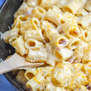 a wooden spoon in a cast iron skillet filled with cheesy riggis pasta with ham