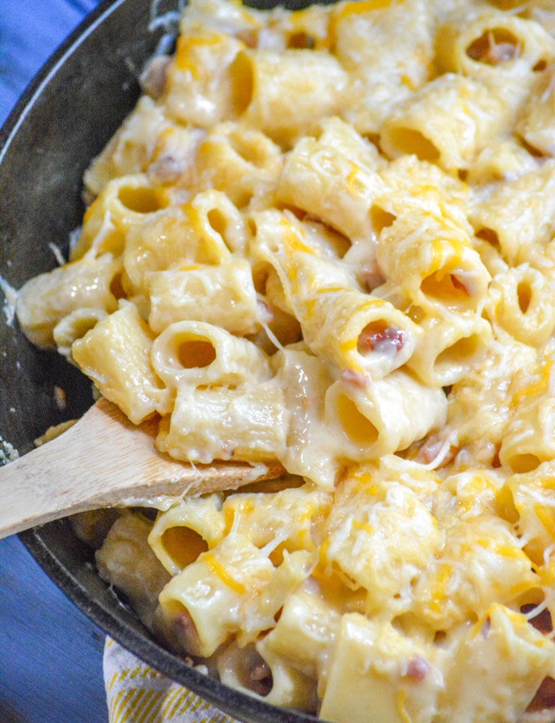 a wooden spoon in a cast iron skillet filled with cheesy riggis pasta with ham