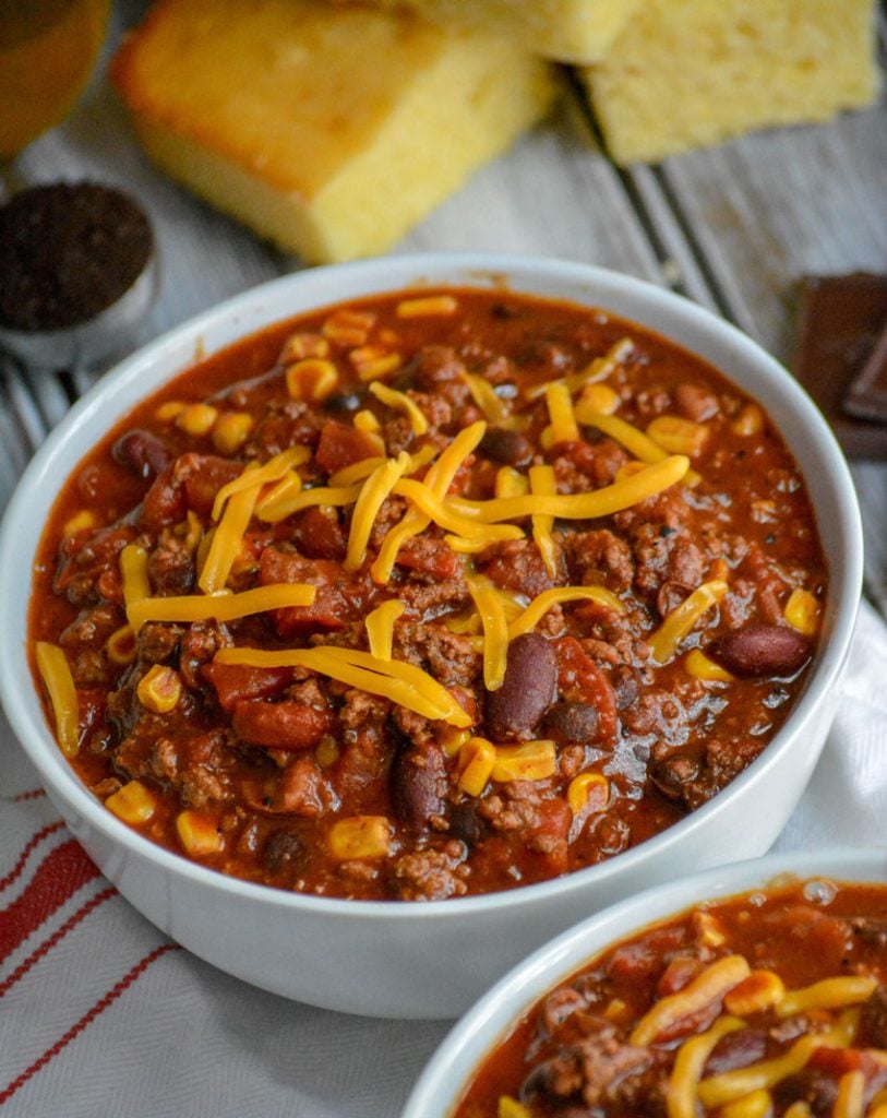 Dad's Award Winning Secret Ingredient Chili served in white bowls, topped with cheddar cheese, and sliced corn bread in the background