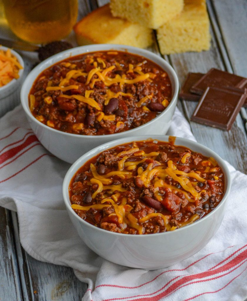 Dad's Award Winning Secret Ingredient Chili served in white bowls, topped with cheddar cheese, and sliced corn bread in the background