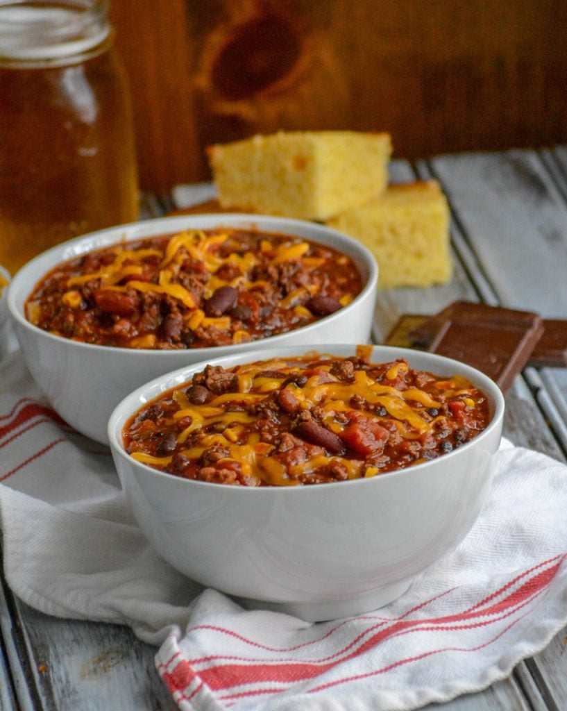 Dad's Award Winning Secret Ingredient Chili served in white bowls, topped with cheddar cheese, and sliced corn bread in the background