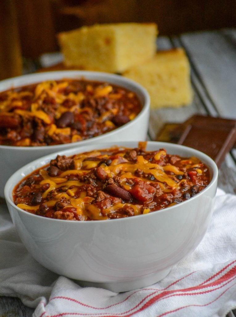 Dad's Award Winning Secret Ingredient Chili served in white bowls, topped with cheddar cheese, and sliced corn bread in the background