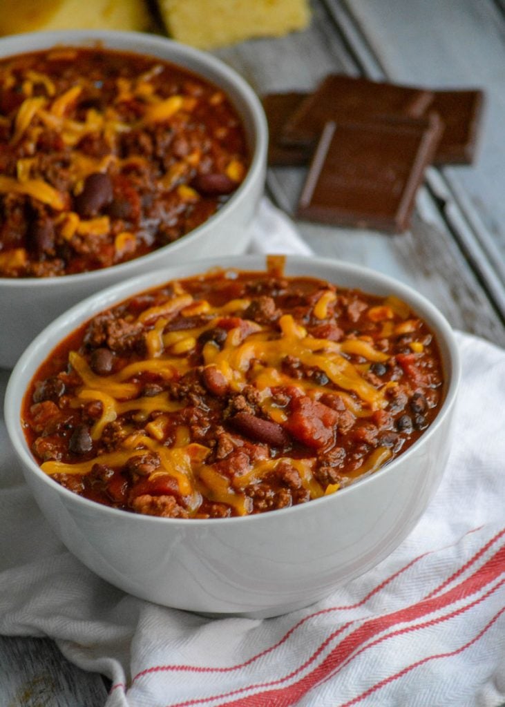 Dad's Award Winning Secret Ingredient Chili served in white bowls, topped with cheddar cheese, and sliced corn bread in the background