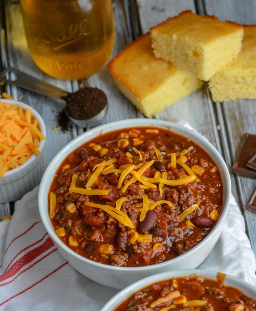 Dad's Award Winning Secret Ingredient Chili served in white bowls, topped with cheddar cheese, and sliced corn bread in the background