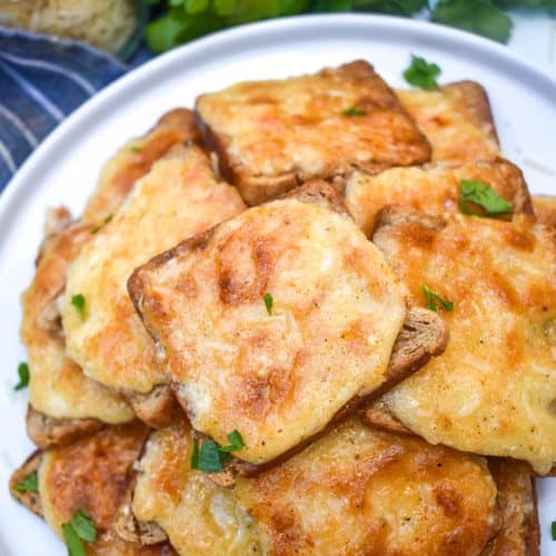 cheesy parmesan rye cocktail bites stacked together on a white plate and topped with a sprinkle of fresh parsley leaves