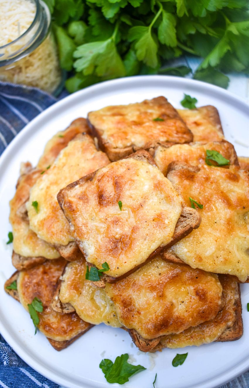 cheesy parmesan rye cocktail bites stacked together on a white plate and topped with a sprinkle of fresh parsley leaves