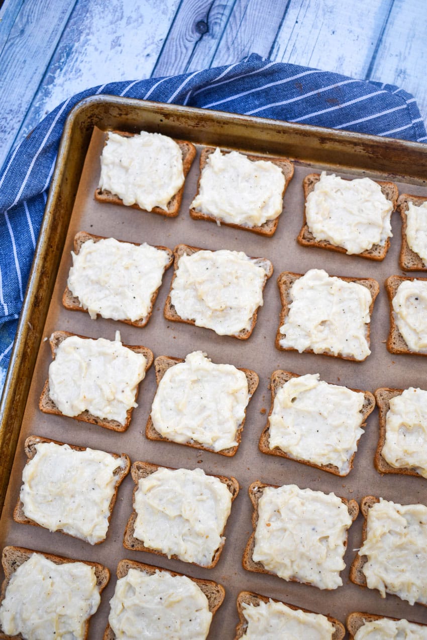 mayonnaise and parmesan cheese spread topped pieces of cocktail rye bread arranged in rows on a metal baking sheet