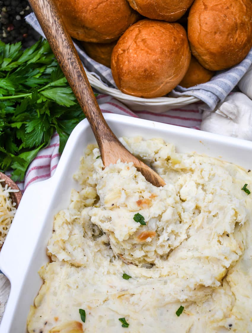 a wooden spoon scooping creamy roasted garlic mashed potatoes out of a white baking dish