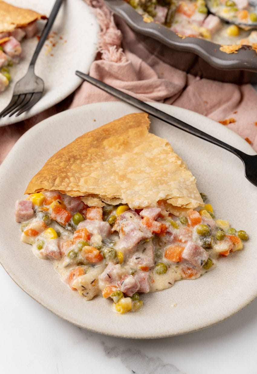 a slice of vegetable cobbler with ham on a white plate with a black fork resting on the side