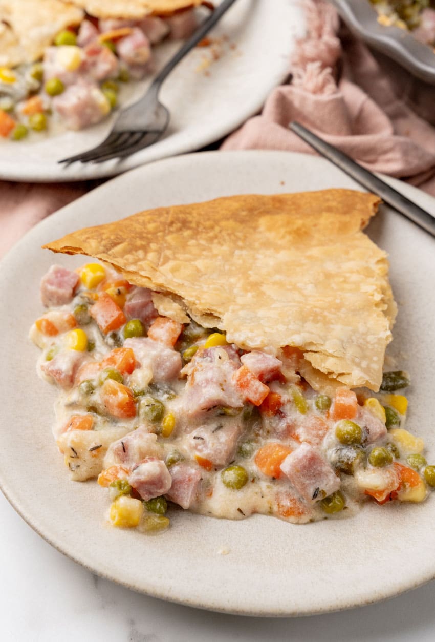 a slice of vegetable cobbler with ham on a white plate with a black fork resting on the side