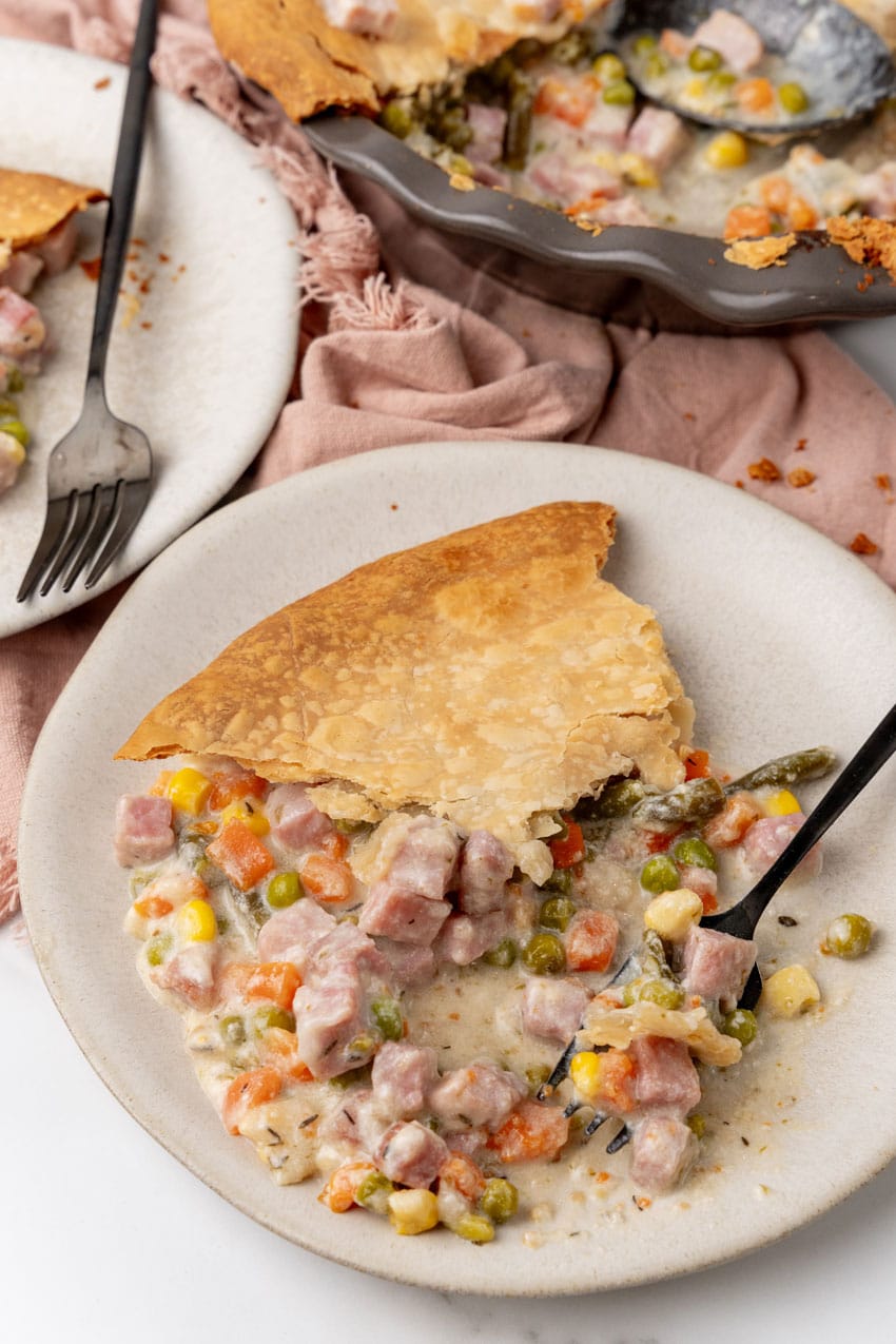 a slice of vegetable cobbler with ham on a white plate with a black fork resting on the side