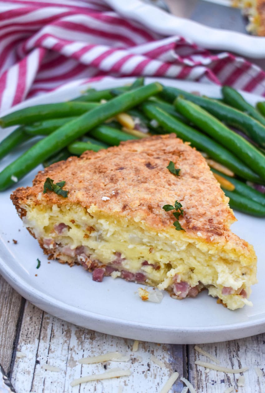 a slice of ham and swiss pie on a white dinner plate next to green bean almondine