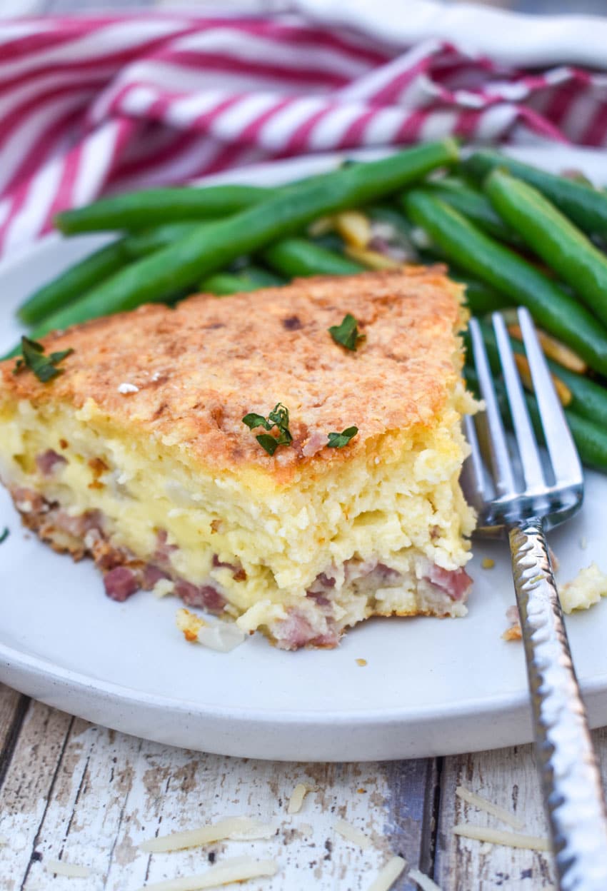 a slice of ham and swiss pie on a white dinner plate next to green bean almondine