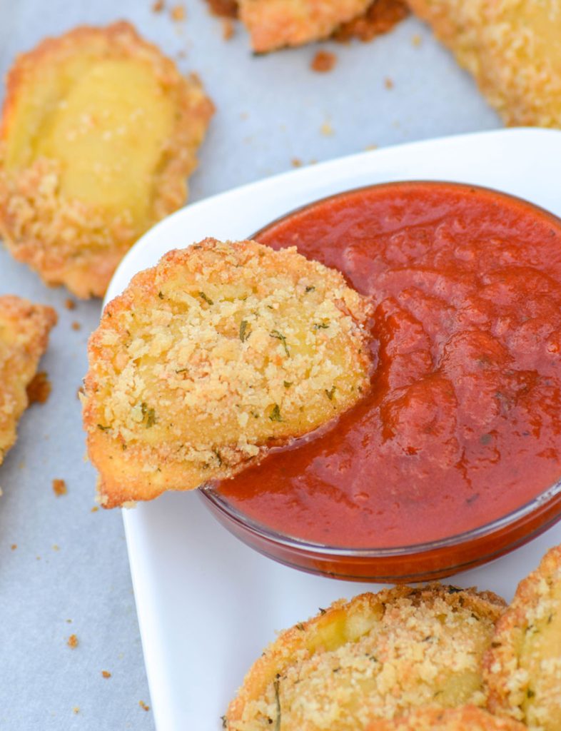 easy oven toasted ravioli recipe showing a piece of toasted ravioli being dipped into a bowl of marinara sauce