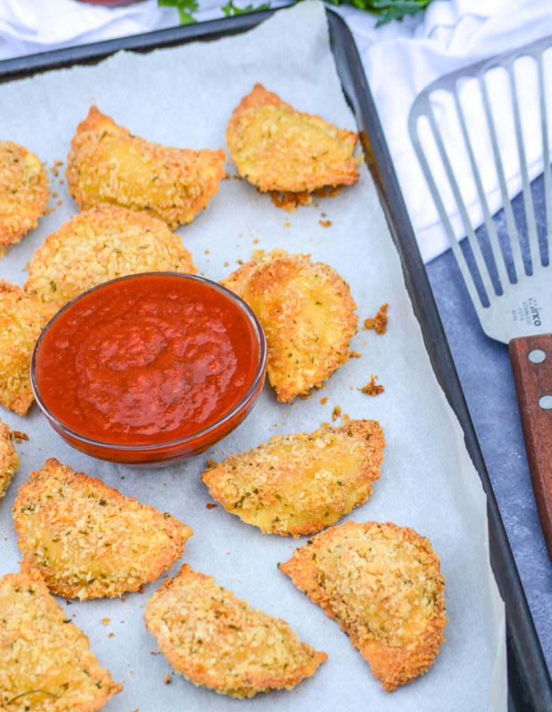 Oven Baked Toasted Ravioli on a parchment covered baking sheet with a bowl of marinara sauce