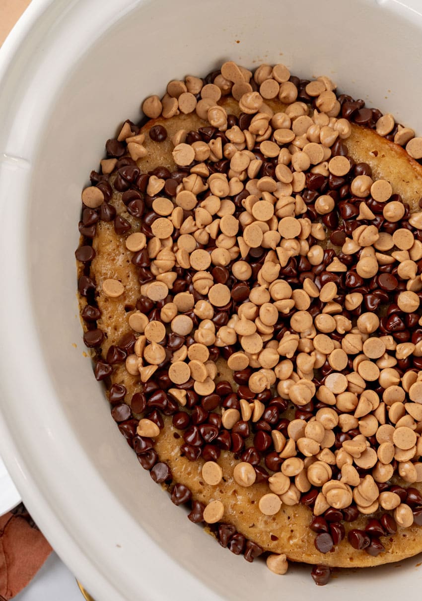 peanut butter chip and chocolate chips overtop a peanut butter banana cake in a white slow cooker