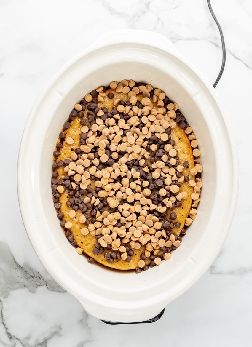 peanut butter chip and chocolate chips overtop a peanut butter banana cake in a white slow cooker