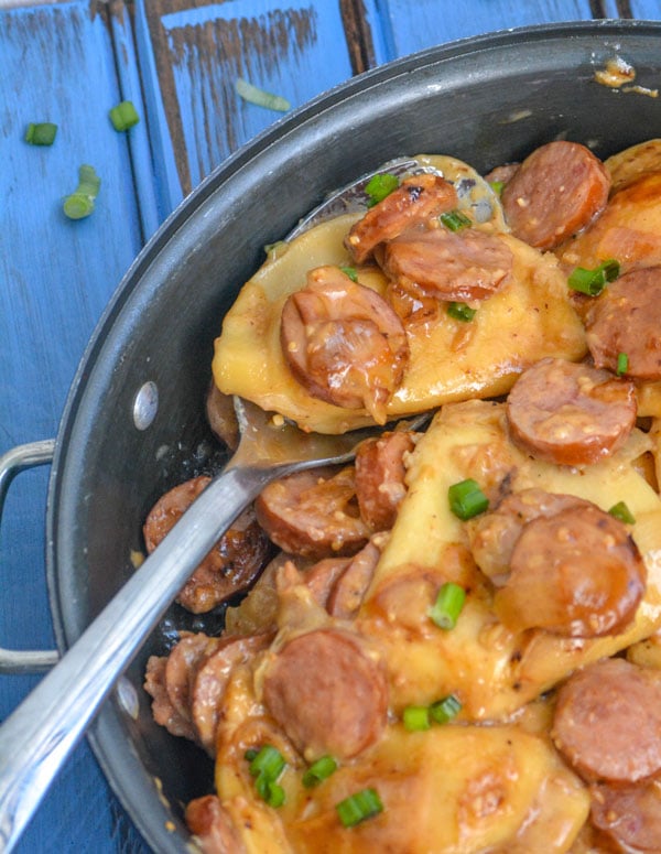 A delicious smoked sausage meal, this one shows sausage and potato stuffed pierogies with caramelized onions and garlic coated with a savory brown sauce. The skillet meal is show being scooped and garnished with freshly sliced green onions.