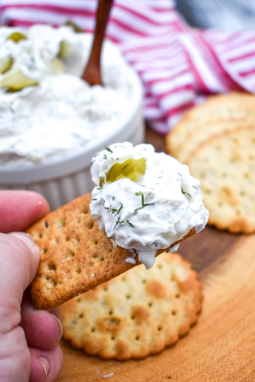 a hand holding up a cracker topped with creamy pickle dip