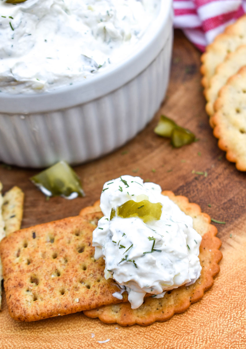 a brown cracker topped with a dollop of a creamy pickle dip recipe resting on a wooden cutting board