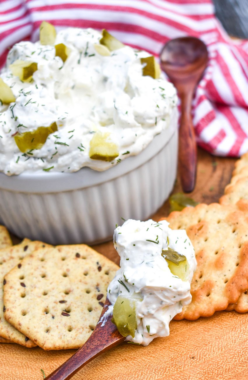 a wooden spoon topped with creamy pickle dip resting on a wooden cutting board surrounded by crackers