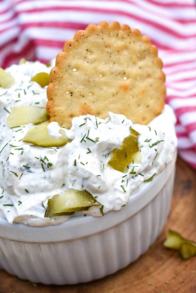 a cracker stuck in a small bowl of dill pickle dip on a wooden cutting board