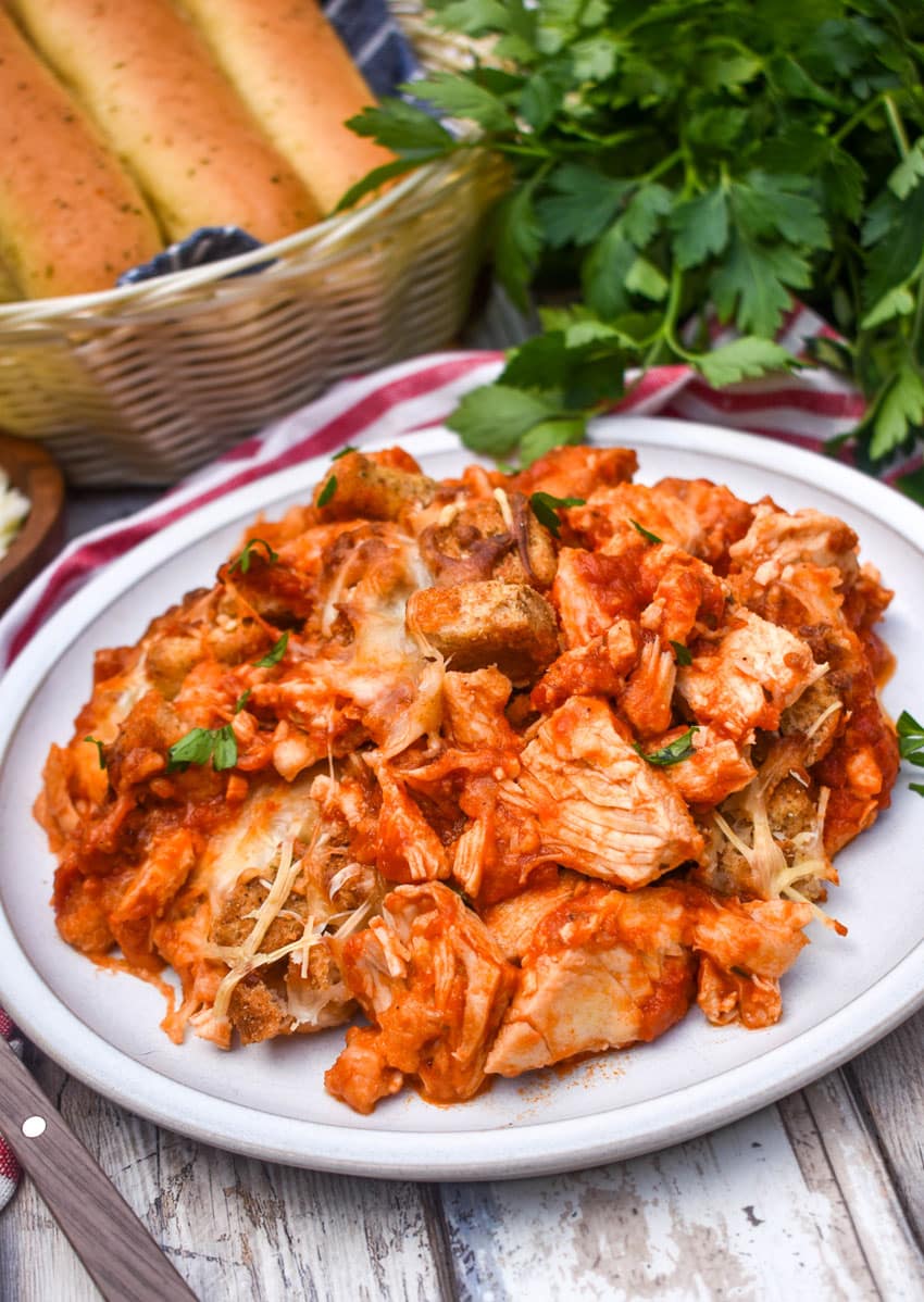 chicken parmesan casserole on a white plate next to fresh green leafy herbs