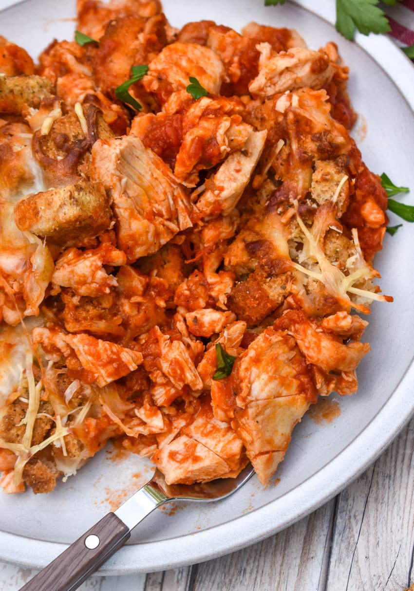chicken parmesan casserole on a white plate next to fresh green leafy herbs