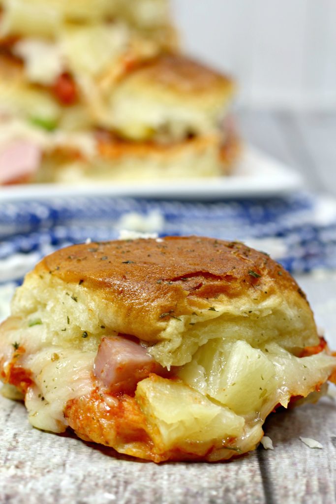 pull apart Hawaiian pizza sliders are piled high on a platter in the background with one single sandwich on a blue cloth in the fore ground