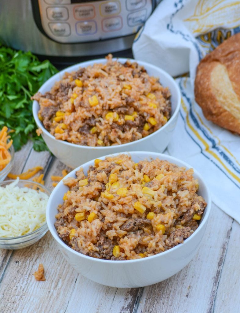 Instant Pot Cheesy Ground Beef & Rice in two white bowls with a browned loaf of crusty bread, an Instant Pot, and bright green Italian parsley in the background