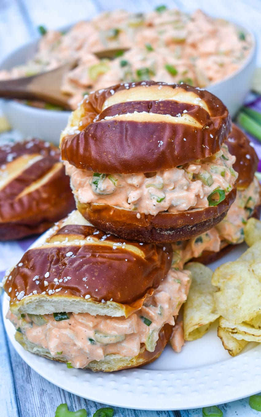 buffalo ranch chicken salad sandwiches on pretzel rolls stacked on a white serving plate with potato chips on the side