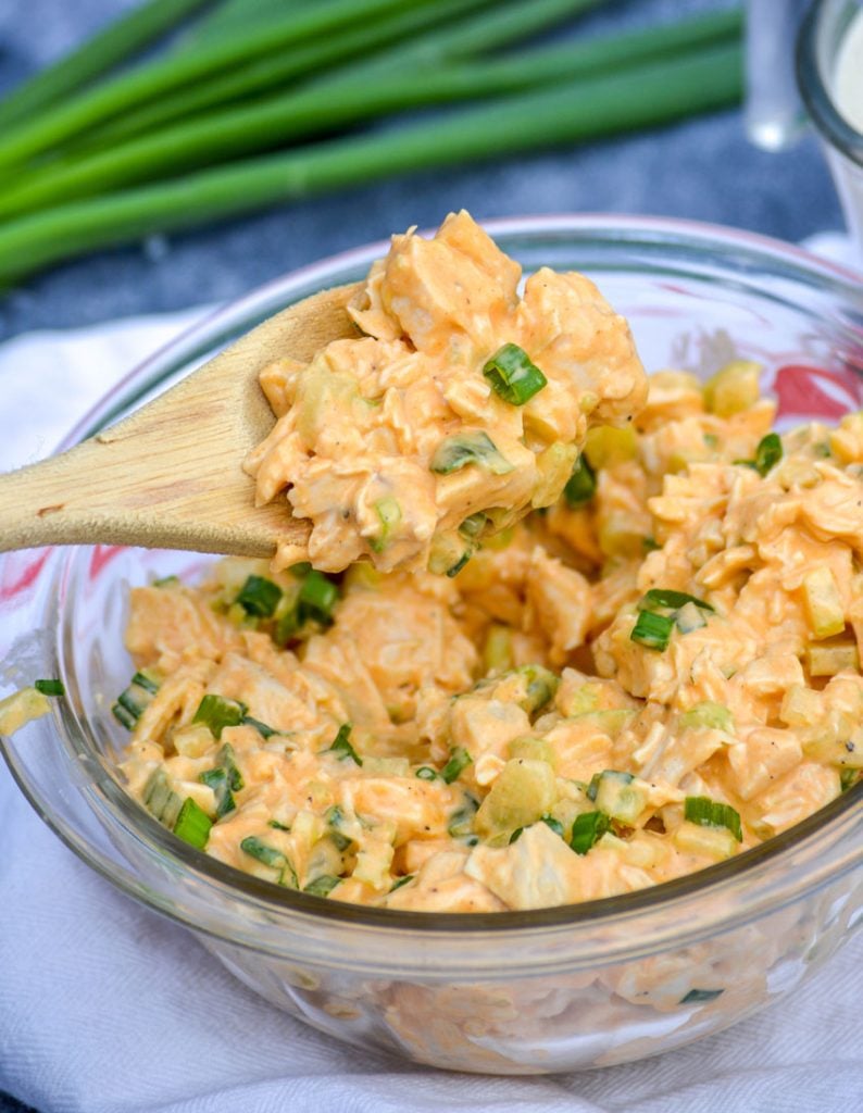 a scoop of creamy buffalo ranch chicken salad is featured on a wooden spoon held above a glass bowl full of the chicken recipe