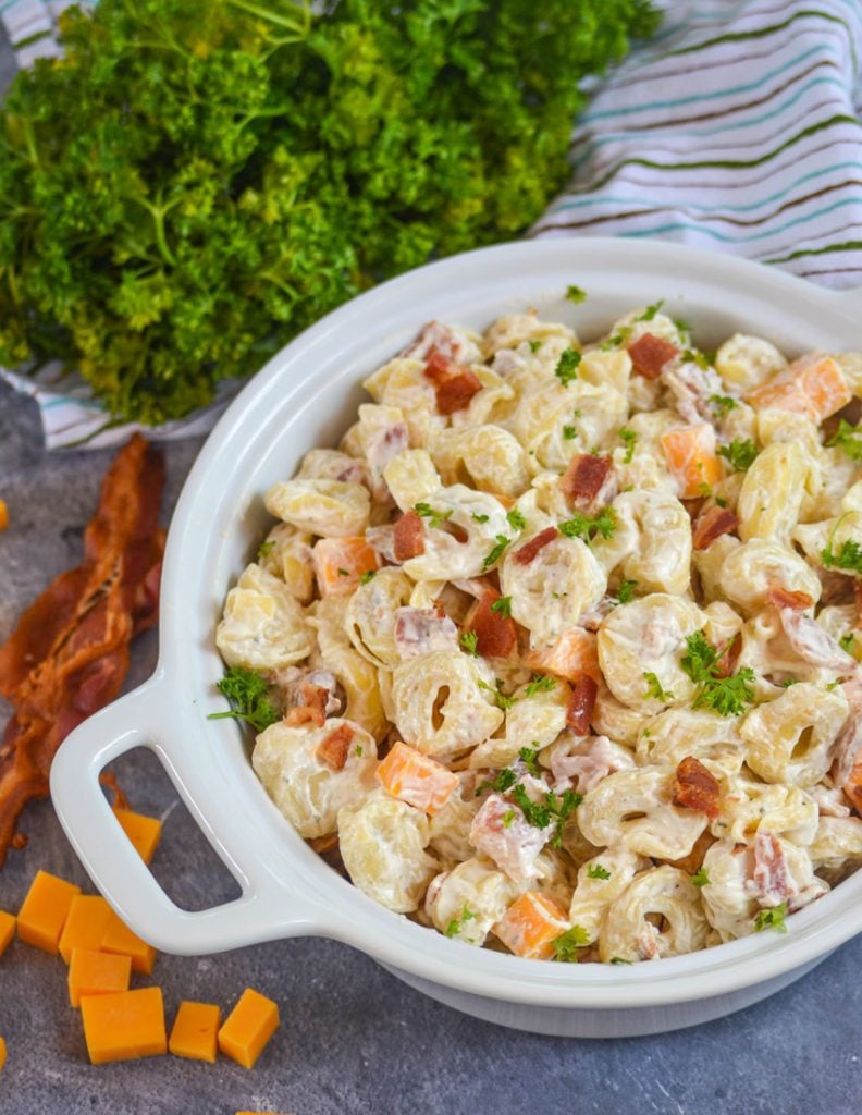 cold tortellini salad with ranch dressing in a white serving bowl on a black marbled counter with crisp slices of bacon and fresh parsley in the background