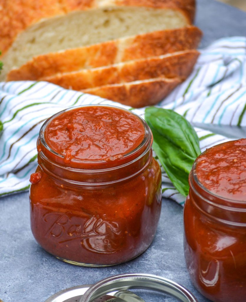 jars of marinara sauce on a gray background with a striped dish towel, basil leaves, and freshly sliced bread