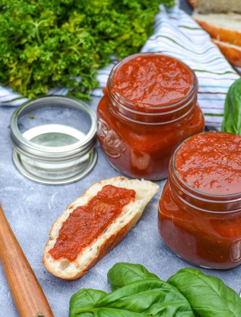 homemade marinara sauce in two glass jars with fresh parsley & basil leaves in the background. A slice of bread is also shown with a spread of marinara sauce.