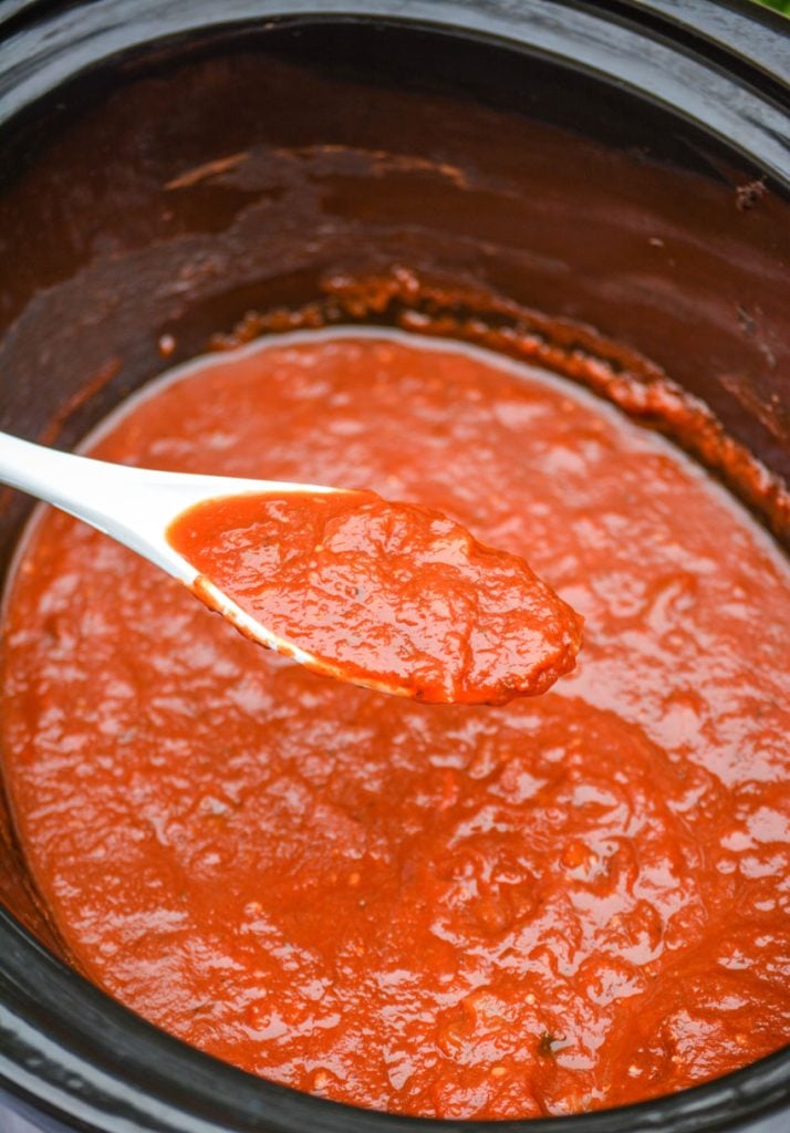 a white spoon holding up some of the red marinara sauce in the black crock of a slow cooker